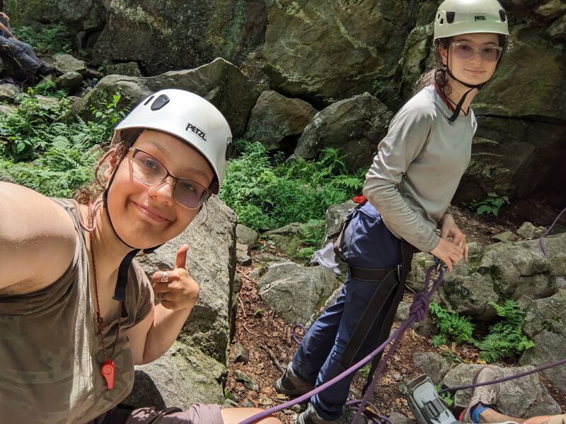 Two young women are posing for a selfie while rock climbing. Both are wearing helmets and climbing gear. The woman taking the selfie is giving a thumbs up. They are surrounded by rocks and greenery. The woman in the background is holding onto a rope, ready to continue climbing.